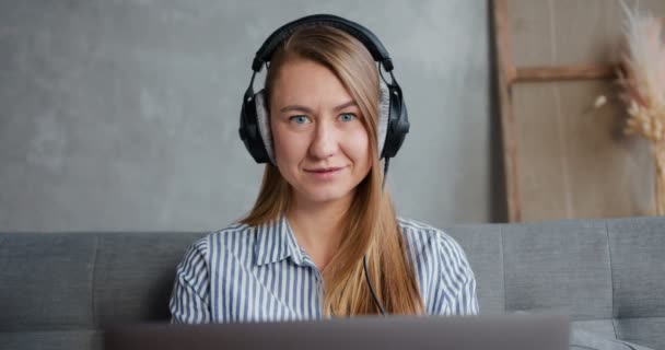 Konzept der Selbstisolierung. Porträt einer glücklichen jungen lächelnden blonden Frau, die Laptop und Kopfhörer benutzt, um von zu Hause aus zu arbeiten. — Stockvideo