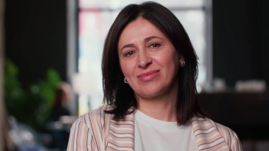 Mutlu, orta yaşlı esmer patron iş kadını modern ışık çalışma ofisinde kameraya gülümsüyor..