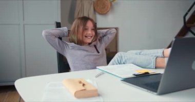 Evde eğitim kavramı, mutlu beyaz kız uzaktan öğretmenle bilgisayar kamerası uygulaması üzerinden çalışıyor, bacakları masanın üstünde.