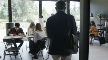 Kamera, genç beyaz iş adamını el dezenfektanı kullanarak modern çok ırklı ofis toplantısına girerken takip ediyor..
