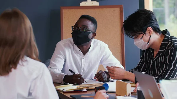 Business meeting with pandemic safety. Happy multiethnic business people work at office table wearing COVID-19 masks.