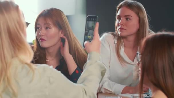 Zwei junge, glückliche multiethnische Geschäftskolleginnen sitzen bei einer Besprechung des Büroteams, während eine andere ein Smartphone-Foto macht. — Stockvideo