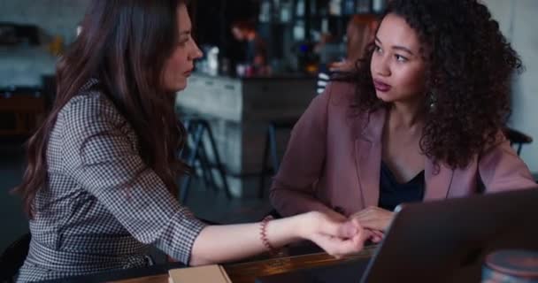 Concepto de entrevista. Dos jóvenes hermosas mujeres multiétnicas hablan usando el ordenador portátil, trabajando juntos en el almuerzo de negocios de la cafetería. — Vídeo de stock