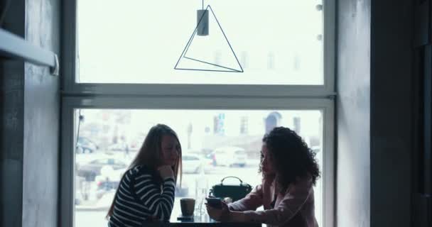 Vista lateral de dos felices mujeres de negocios multiétnicos que se reúnen para el almuerzo, utilizando la aplicación para teléfonos inteligentes en la moderna mesa de la ventana del café de la ciudad. — Vídeo de stock