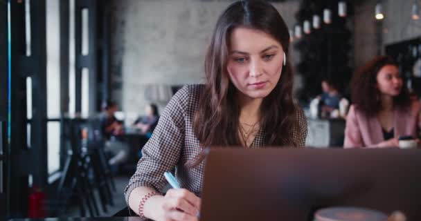 Junge glückliche attraktive nachdenkliche kaukasische Brünette kreative Schriftstellerin macht sich Notizen mit Laptop, um im Café zu arbeiten. — Stockvideo