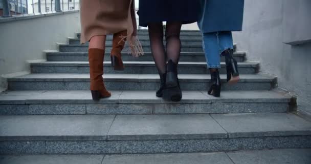 Vue arrière jambes de trois jeunes amies heureuses en manteaux d'automne marchent à l'étage sur la réunion à l'extérieur dans le centre-ville. — Video