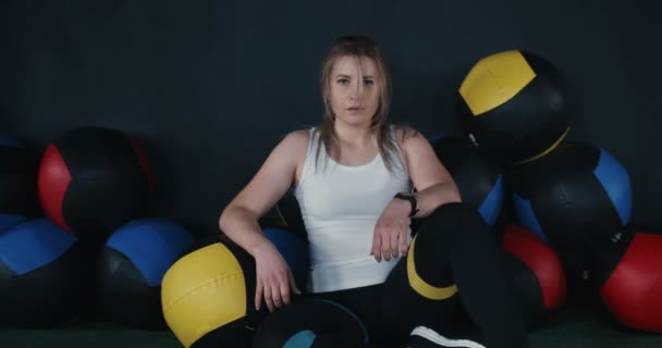 Portrait de belle jeune athlète caucasienne assise sur le sol de la salle de gym parmi les balles de cross fit, épuisée après l'entraînement. — Video