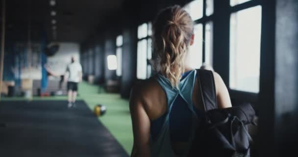 Kamera, sırt çantalı atletik sarışın genç bir kadını büyük modern spor salonuna girerken, spor yapmadan önce arkadaşlarını selamlarken çekiyor.. — Stok video