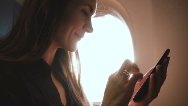 Ατμοσφαιρικό κοντινό πλάνο της νεαρής ευτυχισμένης γυναίκας που χρησιμοποιεί την εφαρμογή για ψώνια smartphone στο κάθισμα παραθύρου του αεροπλάνου, φωτοβολίδα φακού ηλίου. — Αρχείο Βίντεο