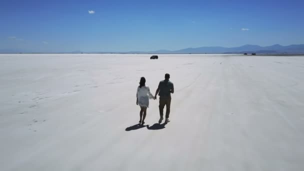 Беспилотник следует за романтической парой, держащейся за руки, направляясь к далекой машине посреди соленого пустынного озера Бонневиль. — стоковое видео
