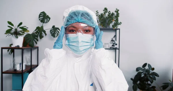 Young multiethnic female doctor wears disposable medical suit and mask, adjusting eyeglasses during pandemic outbreak.