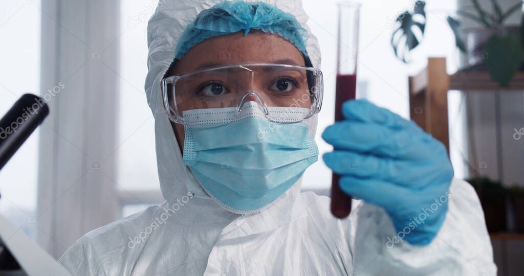 Clinic trials. Young focused mixed race pharma lab doctor scientist in protection suit looking at test tube with blood.