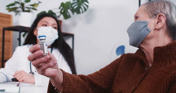 Mixed race pharmacy doctor woman gives flask with vaccine medicine to senior Caucasian patient woman at clinic exam.