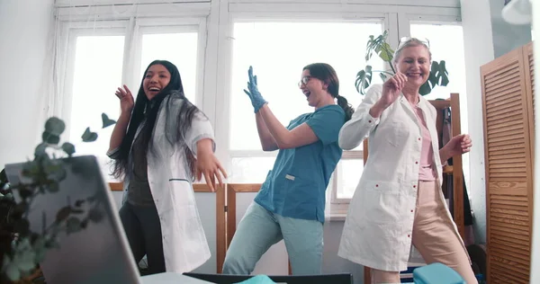 Zoom out three fun happy diverse doctor women dance together celebrating victory over virus pandemic at clinic lab.