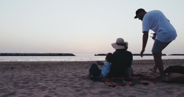 Vista posteriore felice padre si siede insieme a madre e figli a guardare l'alba d'oro epica al mare spiaggia slow motion — Video Stock