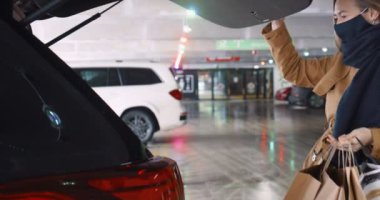 Maskeli genç mutlu sarışın kadın arabanın bagajını açıyor, alışveriş merkezinin otoparkına kağıt poşetler koyuyor..
