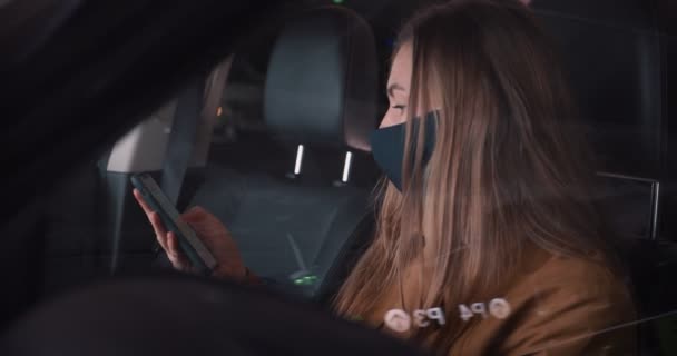 Close-up jonge mooie blonde vrouw in het gezicht masker met behulp van smartphone winkelen app in auto bestuurder stoel op donkere parkeerplaats. — Stockvideo