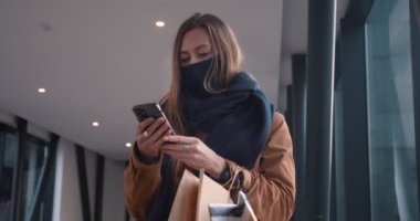 Mutlu, güzel, beyaz maskeli bir kadın alışveriş merkezinin boş koridorunda elinde poşetlerle akıllı telefon kullanıyor..
