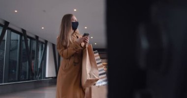 İnternetten güvenli alışveriş. Yüzü maskeli mutlu genç bir kadın alışveriş merkezinin boş koridorunda kağıt torbalarla akıllı telefon kullanıyor..