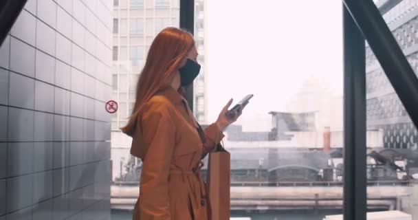 Unterwegs arbeiten. Seitenansicht junge schöne Geschäftsfrau mit Gesichtsmaske mit Handy-App im Einkaufszentrum mit Einkaufstaschen. — Stockvideo