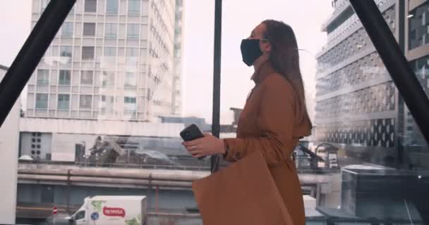 Il concetto di shopping post-pandemico. Giovane bella donna d'affari in maschera cammina lungo la finestra del centro commerciale con sacchetti di carta. — Video Stock