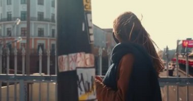 Yan görünüm çekici mutlu blog yazarı kadın akıllı telefon uygulaması kullanarak güneşli şehir köprüsü arka planında yürüyor..