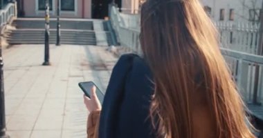 Kamera, güneşli bir günde şehir köprüsünde yürürken akıllı telefon kullanan yüzü maskeli genç sarışın kadını takip ediyor..
