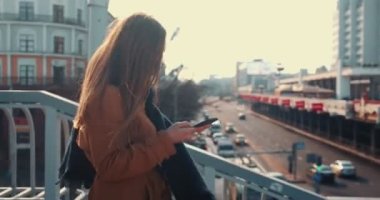 Kamera, neşeli iş kadınını takip ediyor. Yüzünde maske var. Akıllı telefon kullanıyor, güneşli şehir caddesi köprüsü boyunca yürüyor..