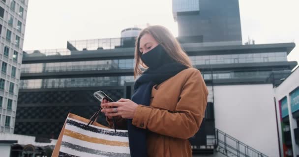 Salgın sırasında güvenli alışveriş. Yüzü maskeli genç ve mutlu bir kadın elinde kesekâğıdı olan akıllı bir telefon kullanıyor.. — Stok video