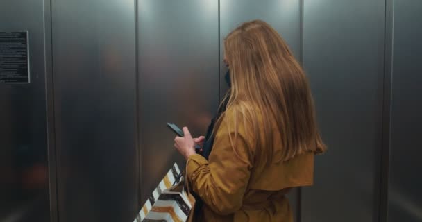Πίσω όψη ευτυχισμένη νεαρή όμορφη γυναίκα των επιχειρήσεων σε μάσκα προσώπου χρησιμοποιώντας το κινητό τηλέφωνο app, αφήνει ασανσέρ με τσάντες ψώνια. — Αρχείο Βίντεο