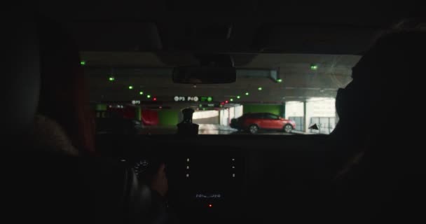 POV shot from inside car. Two women talk while driving personal vehicle along dark parking lot with lights towards exit. — Stock Video