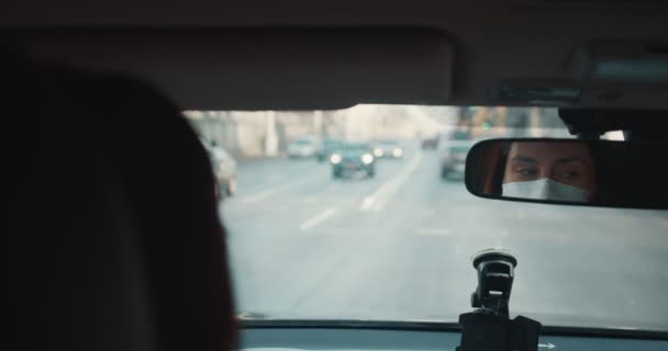 POV achteraanzicht van auto achterbank, ogen van mooie jonge vrouw in gezicht masker rijden langs de straten van de stad reflecteren in spiegel — Stockvideo