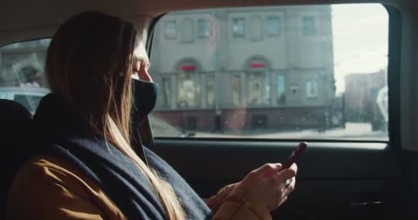 Beautiful thoughtful young blonde passenger woman using smartphone mobile office app in taxi back seat wearing face mask — Stock Video