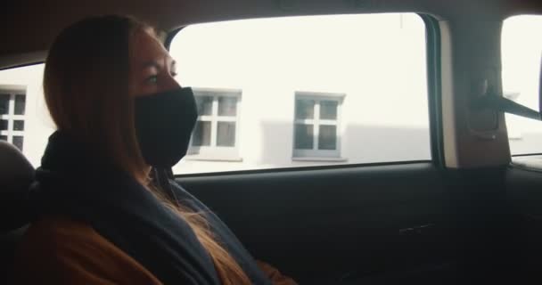 Élet a karantén alatt. Gyönyörű stresszes fiatal szőke nő taxi hátsó ülésen néz ki az ablakon fekete arc maszk. — Stock videók