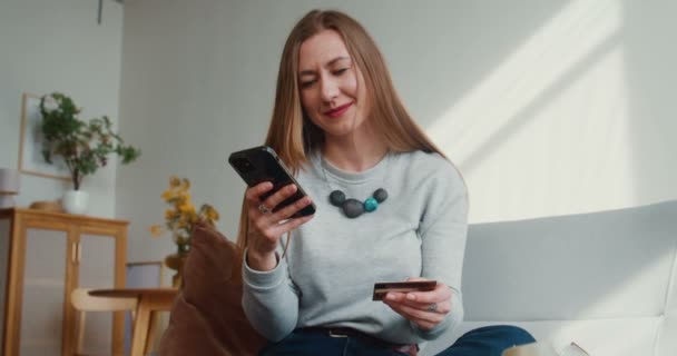 Heyecanlı, mutlu, genç blogcu kadın aydınlık konforlu apartmandaki akıllı telefon ödeme uygulamasına kart numaralarını giriyor.. — Stok video
