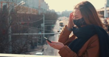 Sosyal medya mobil uygulaması. Maskeli, akıllı telefon kullanan, güneşli şehir manzarasına bakan mutlu yaratıcı genç blog yazarı kadın.