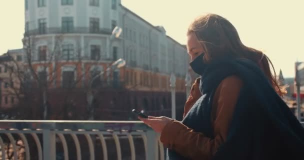 Joven feliz hermosa mujer de negocios en la máscara facial camina a lo largo del puente escénico de la ciudad utilizando la aplicación de teléfono inteligente cámara lenta. — Vídeo de stock