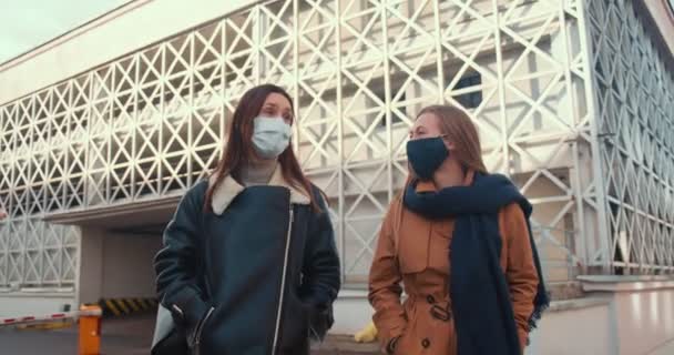 Δύο νεαρές χαρούμενες όμορφες φίλες φορούν ιατρικές μάσκες προσώπου περπατώντας, μιλώντας στο σύγχρονο ηλιόλουστο κτίριο της πόλης — Αρχείο Βίντεο