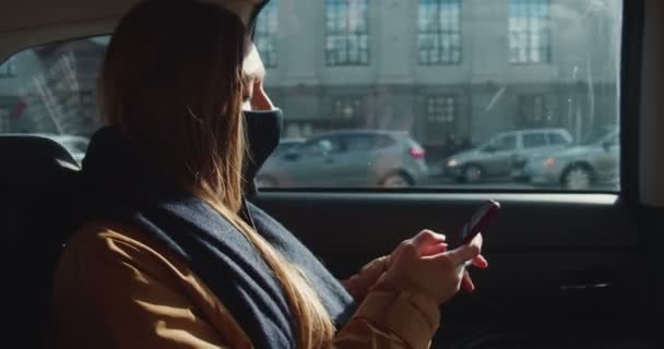 Smartphoneverslaving. Mooie jonge blonde vrouwelijke passagier speelt mobiele game app in taxi achterbank dragen gezichtsmasker. — Stockvideo