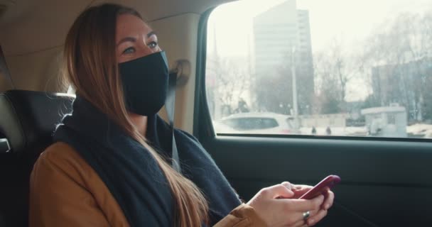 Genç, güzel taksi yolcusu kadın arka koltukta yüz maskesi takarak oturuyor ve blog yazmak için cep telefonu uygulaması kullanıyor.. — Stok video