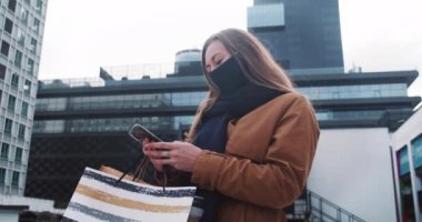 Karantina sonrası alışveriş. Mutlu, genç, güzel, maskeli sarışın kadın elinde kağıt çantalar tutan akıllı telefon kullanıyor.