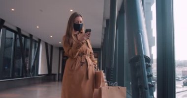Tecritten sonra alışveriş. Mutlu, gülümseyen, maskeli bir kadın alışveriş merkezinin girişinde elinde kesekâğıdıyla akıllı telefon kullanıyor..