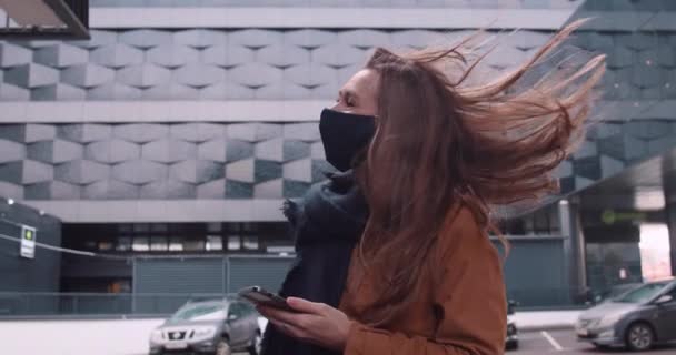 Fiatal szép boldog üzletasszony arc maszk sétál a kamera segítségével okos telefon app nagy futurisztikus épület. — Stock videók