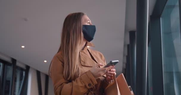Boldog fiatal gyönyörű nő arc maszk néz körül használ okos telefon app pláza folyosón bevásárló papír táskák. — Stock videók