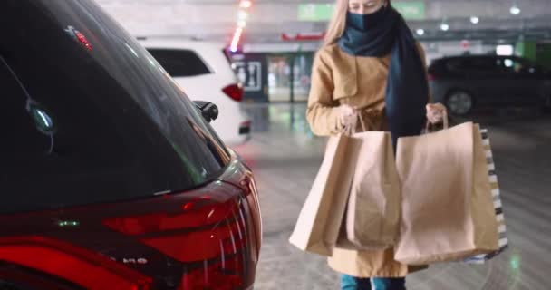 Shopping dopo l'isolamento. Giovane donna bionda felice in maschera apre il bagagliaio dell'auto, mette sacchetti di carta all'interno al parcheggio del centro commerciale. — Video Stock