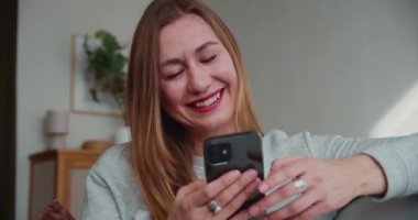 Mobil oyun konsepti. Güzel, mutlu, genç, sarışın bir kadın kanepeye oturup gülümsüyor ve evdeki akıllı telefon uygulamasını kullanarak eğleniyor..