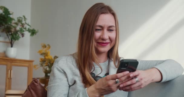 Gyönyörű boldog fiatal mosolygós szőke blogger nő ül a kanapén segítségével smartphone app dolgozni otthonról az önálló elszigeteltség — Stock videók