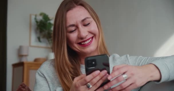 Mobil spel koncept. Vacker glad ung blond kvinna sitter på soffan leenden ha kul med hjälp av smart telefon app hemma. — Stockvideo