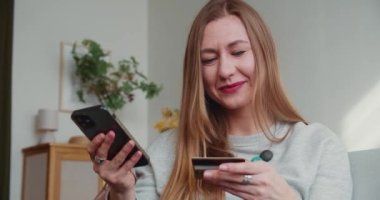 Çevrimiçi ödeme. Güzel sarışın kadın gülümsüyor, kredi kartı numaralarını okuyor, akıllı telefon uygulaması ile eve giriyor..