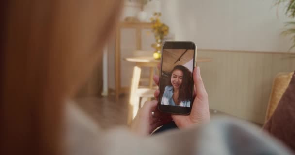 Het concept van een webconferentie. Over schouder uitzicht, blonde vrouw praat met gemengde ras vriend op smartphone scherm bij licht thuis. — Stockvideo